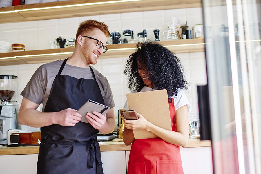 About Our Agency - Man wearing a Blue Smok and a Woman Wearing a Red Smok are Standing Together Checking Inventory at Their Coffee Shop